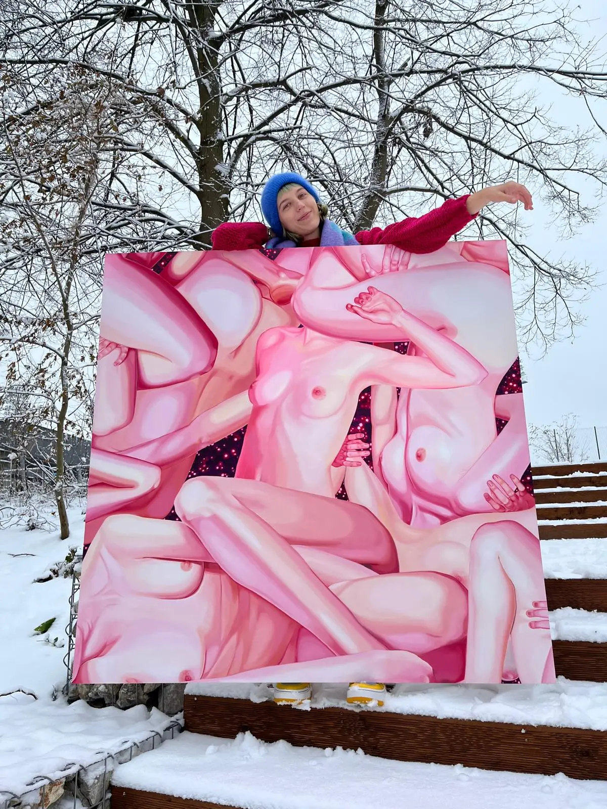 A person standing behind a large painting with pink female nudes, holding it on snow-covered stairs