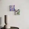 Visualization of two modern surrealist paintings hung on a light wall above a round table with a minimalist sculpture; on the left a painting of a nude figure among blue plants, on the right a painting depicting a female body and a green hand