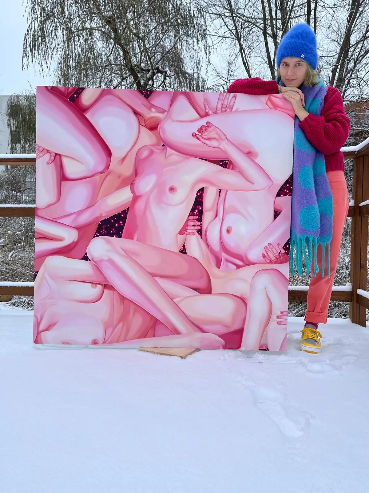 A person holding a painting with pink female nudes, photographed in the snow in a winter outdoor setting