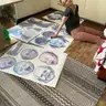 Artystka siedzi na podłodze wśród wyciętych, błękitnych masek z dziecięcymi twarzami