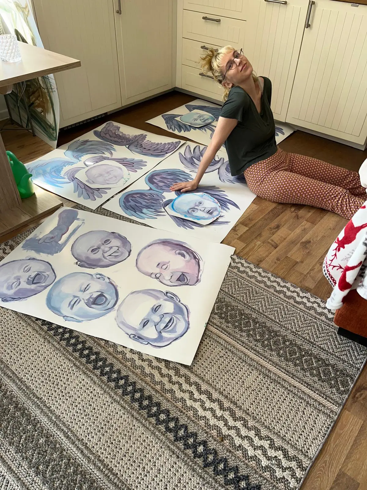 Artystka siedzi na podłodze wśród wyciętych, błękitnych masek z dziecięcymi twarzami