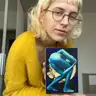 Person in a yellow sweater presenting a small painting with a blue female nude against a moon background